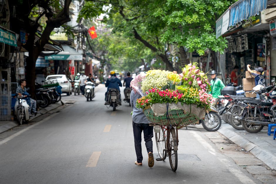 A street scene featuring a flower vendor pushing a bicycle loaded with a vibrant bouquet of fresh flowers, including pink roses, white hydrangeas, and yellow lilies, wrapped in green and beige paper w