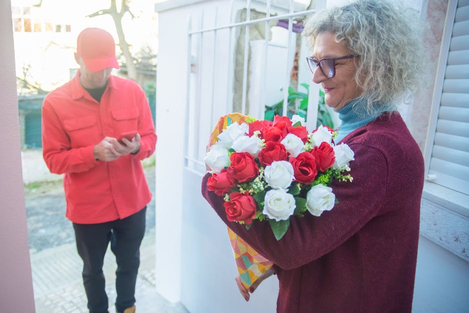 A woman with curly gray hair and glasses is holding a colorful bouquet of fresh red and white roses wrapped in bright, patterned paper, standing near a white door with shutters in a bright, well-lit e
