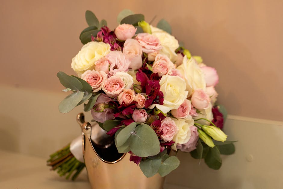 A floral arrangement featuring a bouquet of fresh pale pink roses, white roses, and deep burgundy alstroemeria, accented by dusty eucalyptus leaves arranged in a compact, rounded style. The bouquet is