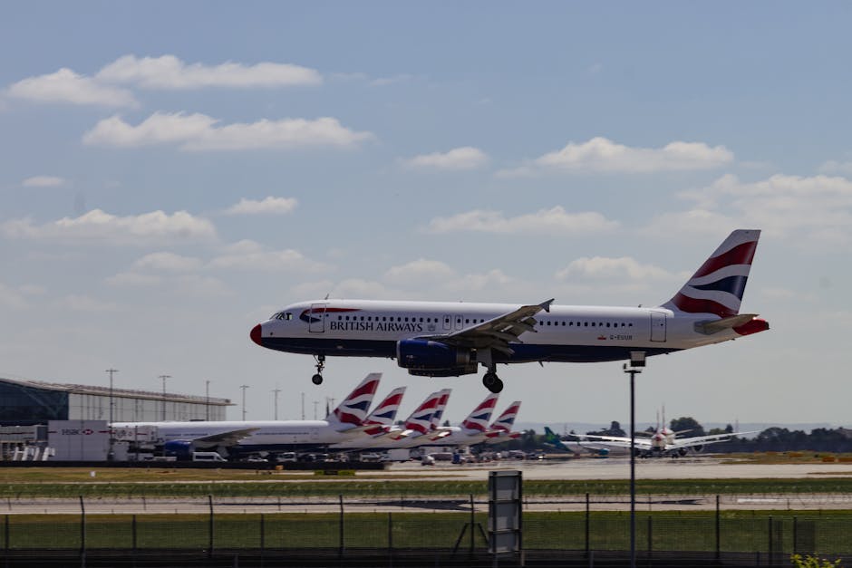 An aircraft painted with the British Airways livery is captured mid-flight as it approaches an airport runway, with several other British Airways airplanes parked on the tarmac below. The background f