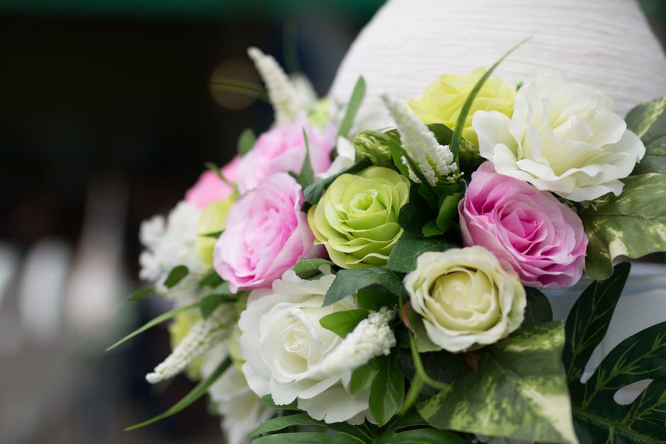 A close-up of a floral arrangement featuring an assortment of fresh roses in soft pink, ivory, and pale green hues, complemented by white lavender-like flowers and lush green foliage. The bouquet is d
