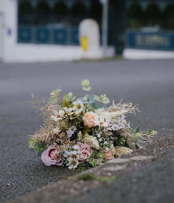 A delicate floral bouquet placed on an asphalt street beside a curb, featuring soft pink roses, white daisies, and small white filler flowers, accented with pale green eucalyptus leaves and wispy drie