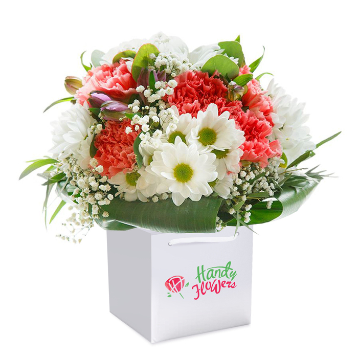 Brought to life in a sleek white gift bag, this vibrant floral bouquet is a celebration of Harmondsworth's colour and charm, expertly arranged to delight both giver and receiver. Lush clusters of coral, peach, and vibrant red carnations command attention with their abundant, frilled petals, radiating both warmth and sophistication. These are interwoven with immaculate white daisies bearing striking yellow-green eyes-cheerful bursts reminiscent of garden blooms along the village's quiet streets. Lending softness, sprigs of dainty baby's breath (gypsophila) create a light, ethereal effect, while on one side, buds tinged with purple promise future splendour. Deep green, glossy foliage wraps the bouquet, offering contrast and providing a supportive, natural enclosure. Each element is perfectly placed, ensuring a full, rounded silhouette-emphasising freshness, balance, and abundance. Sturdy ribbon handles emerge from the top of the bag, inviting an easy and elegant handoff perfect for same-day delivery to homes near Harmondsworth Village Green. Photographed against a snow-white background, the details pop: the intricate play of texture, the lively colour palette, and the impression of a bouquet meant to mark occasions both grand and intimate. This arrangement stands as an irresistible token of affection for birthdays, anniversaries, or simply to share a piece of village cheer.