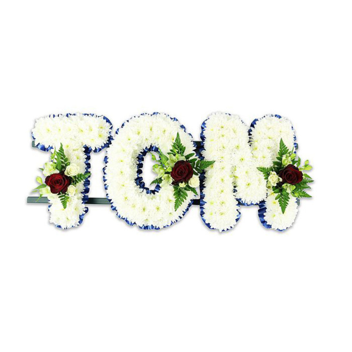 A personalised sympathy piece presents the name 'TOM' in substantial, pillow-like letters entirely cloaked in pure white chrysanthemums, each tiny bloom placed to form a smooth, cloud-like surface that reads as one continuous field of white. The edges of each letter are crisply defined with a vibrant royal blue ribbon, the satin trim providing a composed contrast that outlines the type and keeps the tribute visually tidy against the stark white background. Each of the three letters is embellished with a measured floral spray: a solitary, deep red rose anchors the cluster, chosen as the focal signifier of love and remembrance, while surrounding ivory spray roses and sprays of delicate fern bring texture, subtle fragrance and a soft green accent. The composition reveals careful handcrafting - blooms graded for size, fillers placed to create depth, and the ribbon tucked neatly so the overall effect remains formal and respectful. A dark green or black metal framework is visible beneath the base, a discreet reminder of the structural support needed for standing displays at services. The lighting in the image is even and soft, throwing gentle shadows that emphasise the letters' three-dimensionality and the chrysanthemum's individual petals. For memorials in and around Harmondsworth - whether at the parish church, a funeral home off Bath Road or a chapel close to Heathrow - this style communicates tastefully curated sympathy: restrained, tactile and designed to allow mourners to see a loved one's name presented with calm dignity and floral skill.