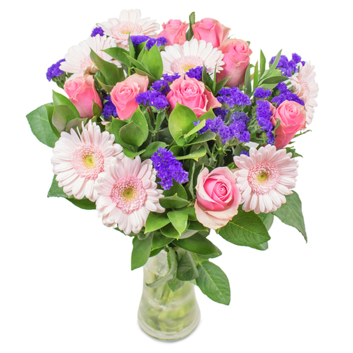 A striking bouquet-arranged by hand in Harmondsworth-showcases around eight blush-toned roses, each unfolding layers of delicate pink petals in the classic spiral that signifies romance and gratitude. Softly fringed gerbera daisies in a paler pink are interspersed throughout, their cheerful, wide faces radiating warmth. Punctuating the display are clusters of deep lavender statice, lending dramatic texture and colour depth, much like the wildflowers along the banks of the River Colne. A base of lush, emerald-green foliage anchors the arrangement, and the stems rest in a cylinder of clear glass, water sparkling within. Visible from all angles, this bouquet seems to pulse with the freshness of an early morning walk through Harmondsworth's green spaces. It's the kind of gift well-suited to birthdays, anniversaries, or simply as an uplifting surprise-an arrangement that merges artistry and local charm in every carefully placed bloom.