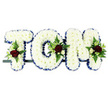 This image captures a personalised funeral flower arrangement that spells out the name TOM in large, three-dimensional capital letters, ideal for a memorial or graveside tribute in Harmondsworth. Every letter is densely filled with pristine white chrysanthemums, their small, tightly packed blooms forming a soft yet clearly defined surface. Around the edge of each letter, a rich royal blue ribbon is neatly pleated, tracing the outline and adding a calm, dignified contrast to the white petals. At carefully chosen points-a corner of the T, the side of the O, and the upper section of the M-small sprays of feature flowers emerge from the chrysanthemums. These include deep crimson red roses in full bloom, creamy white and soft white spray roses, and slim, tubular white flowers that resemble freesia, adding gentle height and elegance. Lush green foliage, including fern and other mixed leaves, fans out behind the roses, creating depth and a natural, garden-like feel. The letters are aligned horizontally on a narrow dark frame, ready to be placed on a coffin, along the front of the dais at a crematorium, or near a photograph at a remembrance gathering. A plain white background keeps the focus on the tribute's colours and textures, emphasising the contrast between pure white, royal blue, deep red, and fresh green. The arrangement conveys a sense of respect and personal connection, a custom floral memorial for someone named Tom, thoughtfully designed by local florists serving the Harmondsworth community who understand the tenderness needed for final farewells.
