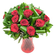 A lush, rounded bouquet of a dozen or more deep crimson roses fills the frame, each bloom opened to a full, velvety perfection. The petals spiral tightly from a dense centre, their wine-dark red catching a soft, even light that emphasizes velvet texture and delicate petal edges. Around the rose heads a generous chorus of dark green foliage-think glossy Ruscus or Salal-forms a verdant backdrop, while broader, emerald-green leaves, folded and rolled like polished ribbons, are tucked into the composition to create sculpted loops and graceful arcs. Those architectural leaf curls lend a contemporary edge and visually cradle the roses, adding height and structure without stealing the intimate, romantic focus. The whole composition sits in a tall, slender ruby-red glass vase with a subtle ribbed or twisted surface; light travels down the vase's vertical lines, picking out deeper burgundy highlights that mirror the rose tones and lengthen the silhouette. Photographed against a crisp white background, the arrangement's colours pop with clarity, the reds and greens defined by bright, even illumination. Up close you can almost sense the soft fragrance-classic, warm, quietly floral-inviting thoughts of anniversaries, Valentine's moments, or a thoughtful celebration. As a Harmondsworth-grown design influence, this bouquet feels equally at home on a dining table in the village or as a graceful gift delivered to a nearby Heathrow address, offering both timeless romance and contemporary floral craftsmanship.