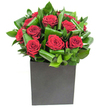 This elegantly modern arrangement reads like a quiet love letter: a tight cluster of rich, velvety red roses sits at the heart of the design, each bloom opening in layered spirals that catch the light with a soft, velour sheen. Broad, glossy green leaves have been lovingly curled and interlaced around the roses, their sculptural loops creating a rhythmic, almost architectural pattern that steadies the crimson intensity and adds a cool, tropical freshness to the composition. The flowers are set squarely in a matte black cube that anchors the display with contemporary confidence; its smooth, understated surface contrasts with the dewy highlights on each petal and the faint waxiness of the foliage. The studio lighting is bright and consistent, revealing luminous reds and jewel-like greens and throwing small, gentle reflections across the black vessel as if the bouquet itself were a carefully wrapped present. Up close, the textural contrast is clear: silk-soft petals beside firm, glossy leaves, and beneath it all a clean, uplifting scent of classic rose lifted by green, herbaceous notes. As a local florist familiar with the lanes around Harmondsworth and the quiet charm of the village green near the Great Barn, I imagine this piece brightening an intimate anniversary dinner, accenting a contemporary office near Bath Road, or waiting as a thoughtful arrival for a romantic airport reunion. Its measured drama and refined silhouette make it a lasting visual statement - confident, tender, and utterly present in the moment.